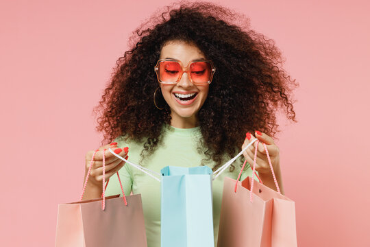 Exultant happy young curly latin woman 20s wears mint t-shirt sunglasses holding looking into package bags with purchases after shopping isolated on plain pastel light pink background studio portrait.