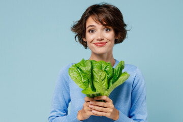 Young smiling cheerful caucasian woman 20s wearing casual sweater look camera hold green lettuce leaves isolated on plain pastel light blue background studio portrait. People lifestyle food concept.