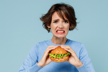 Young sad irritated stressed woman 20s wearing casual sweater look camera biting eating burger isolated on plain pastel light blue color background studio portrait. People lifestyle junk food concept.