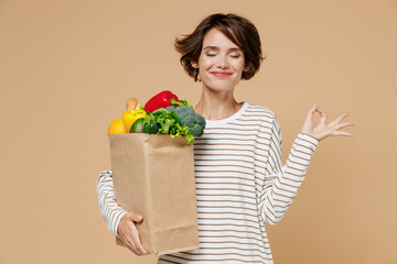 Young woman in casual clothes hold paper bag with vegetables hold spreading hands in yoga om aum...