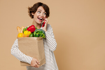 Young smiling woman 20s in casual clothes hold paper bag with vegetables speak talk on mobile cell...