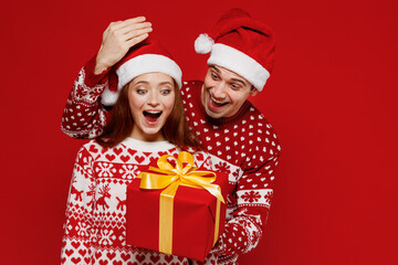 Young couple friends two man woman in sweater hat give red present box with gift ribbon bow open eyes isolated on plain red background. Happy New Year 2022 celebration merry ho x-mas holiday concept.