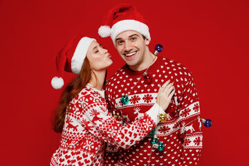 Young lovely couple friends two man woman in sweater hat hold colorful garland kiss isolated on plain red background studio portrait. Happy New Year 2022 celebration merry ho x-mas holiday concept.