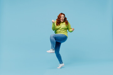 Full body overjoyed young chubby overweight plus size big fat fit woman wear green sweater do winner gesture raise up leg isolated on plain blue background studio portrait. People lifestyle concept.