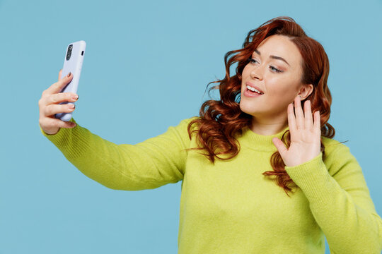 Young smiling happy chubby overweight plus size big fat fit woman wearing green sweater doing selfie shot on mobile cell phone waving hand isolated on plain blue background. People lifestyle concept.
