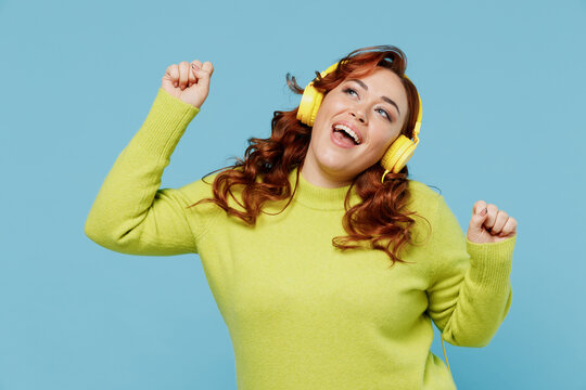 Young smiling cheerful happy chubby overweight plus size big fat fit woman wear green sweater headphones listen to music dancing isolated on plain blue background studio. People lifestyle concept