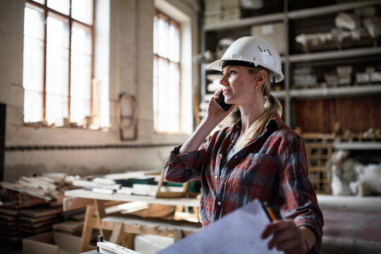 Female engineer holding blueprints and making phone call indoors in carpentry workshop.