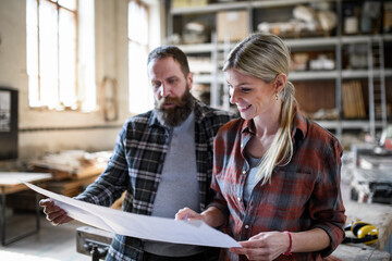 Two carpenters man and woman looking at blueprints indoors in carpentery workshop.