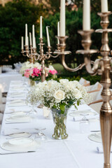 Fototapeta na wymiar bouquet of white roses and gypsophila on a table decorated with large candelabra on a white tablecloth with cutlery on the background of the greenery of garden