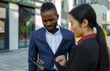 Colleagues African American man with stubble in formal suit and Asian young woman with long brunette hair check work using tablet PC