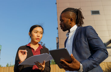 Colleagues black man with curly ponytail in navy suit and Asian young brunette woman discuss project holding tablet and diagrams near office