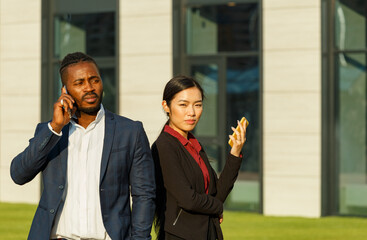 Business partners African American man in navy suit and young Asian brunette long-haired woman solve work issues using phones near office lawn - 471980053