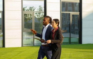 Colleagues African American man in suit and Asian young brunette long-haired woman stroll near office solving work issues with tablet - 471980055