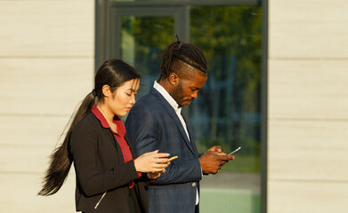 Business partners African American man in suit and Asian long-haired woman surf social nets using phones and strolling near office - 471980059