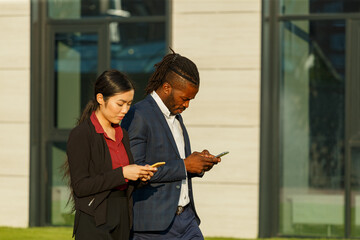 Business partners African American man in suit and Asian long-haired woman surf social nets using phones and strolling near office - 471980061