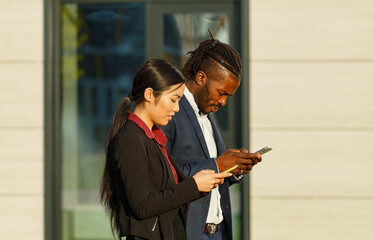 Business partners African American man in suit and Asian long-haired woman surf social nets using phones and strolling near office - 471980062