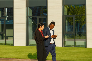 Business partners African American man in suit and Asian long-haired woman surf social nets using phones and strolling near office - 471980063