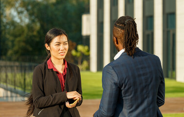 Partners in business African American man in suit and Asian brunette long-haired woman communicate standing near office building at sunset - 471980065