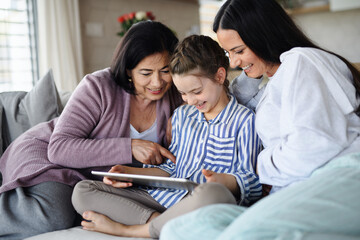 Fototapeta premium Happy small girl with mother and grandmother indoors at home, using tablet.
