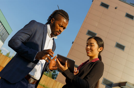 African American Man In Navy Suit And Asian Young Brunette Woman Discuss Work Issues Using Tablet And Standing Near Office Low Angle Shot