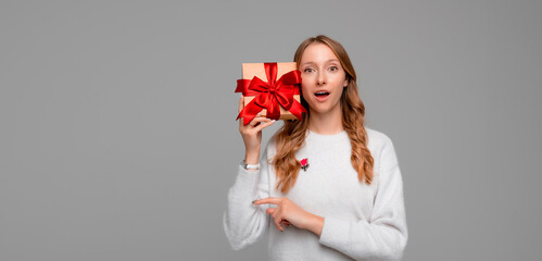 Portrait of amazing young blonde woman listening what rattles in gift box, standing over gray background. New Year Women's Day birthday holiday concept. Mock up copy space
