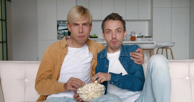 Two LGBTQ men watching horror movie film on TV and Eating Popcorn at Home. They are shocked by the horror scene