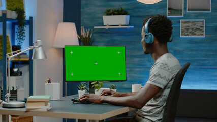 Entrepreneur working on computer with horizontal green screen. Man wearing headphones, using chroma key with isolated mockup template and background. Person with virtual display