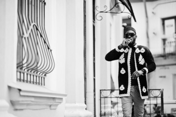 Mega stylish african man in traditional jacket pose. Fashionable black guy in hat and sunglasses with cigar in hand.