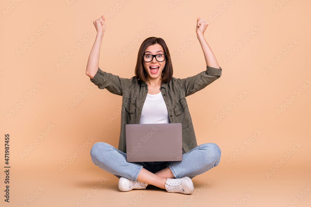 Wall mural photo of lady sit floor hold laptop rejoice raise fists wear glasses khaki shirt isolated beige colo