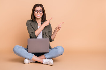 Photo of cheerful lady sit floor hold pc indicate finger empty space sale wear khaki shirt isolated beige color background
