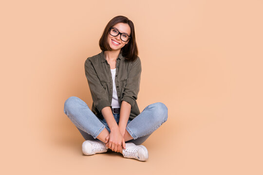Photo of cheerful lovely lady sit floor cuddle legs beaming smile wear specs khaki shirt isolated beige color background