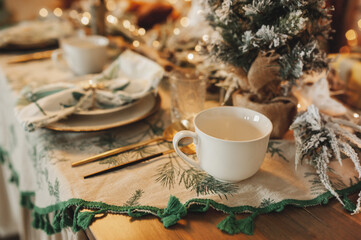white ceramic mug on chic Christmas table setting, beige and wooden tones, candles, cotton and garlands on the background of bokeh and Christmas interior, selective focus