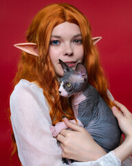Portrait of young female cosplay elf, holding adorable bicolor Sphinx kitten in hands. Elf with beautiful wavy red long hair, in summer white dress, smiling and looking at camera. Red background.