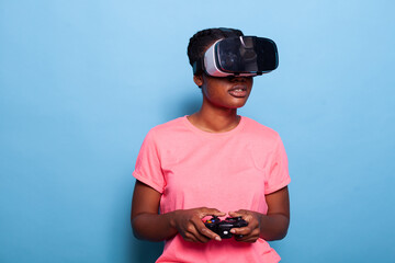 Portrait of african american teenager gamer wearing virtual reality headset playing entertainment video games using gaming joystick standing in studio with blue background. Videogames concept