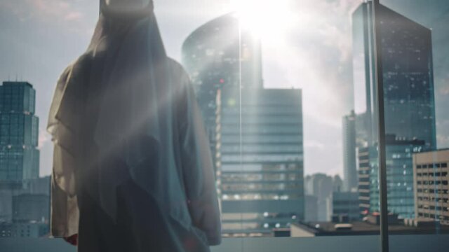 Successful Muslim Businessman in Traditional White Kandura Walking in His Modern Office Looking out of the Window on Big City with Skyscrapers. Successful Saudi, Emirati, Arab Businessman Concept.
