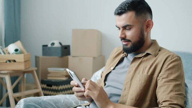 Cheerful Middle Eastern man is using smartphone touching screen sitting in new apartment with cardboard boxes enjoying communication. Gadgets and relocation concept.