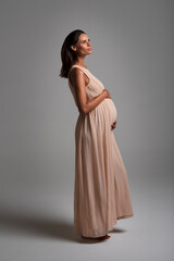 Young beautiful pregnant woman standing on the floor wearing a long pink dress vertical view grey background. High quality photo