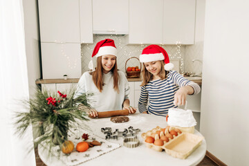 Fototapeta premium Two charming sisters in Santa Claus hats bake cookies for Christmas in the kitchen. Christmas cooking, preparation for the holiday, Christmas cookies