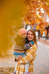 Portrait of happy and beautiful mother with her daughter in her arms among autumn leaves on city street. Fall colors.