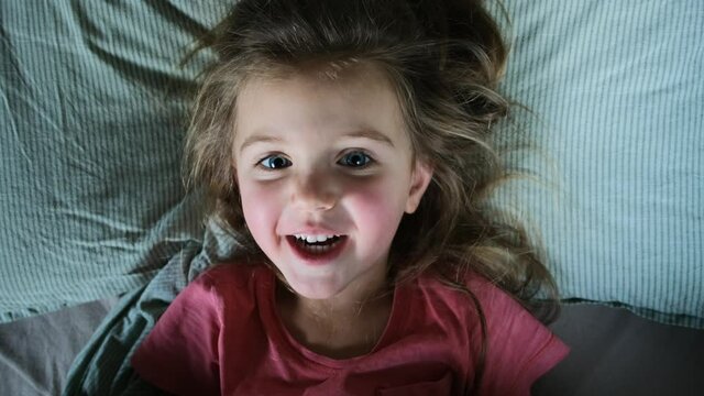 Portrait of upset sleepy little girl looking at camera with sadness amazed frustration in her eyes, lonely kid cute lady lying back in bed her face close-up. Child starts to have fun and laugh