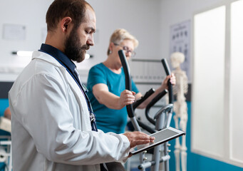 Naklejka na ściany i meble Senior patient using stationary bicycle for physical recovery while medic looking at tablet for healthcare. Woman with injury doing exercise activity for fitness and physiotherapy