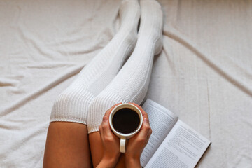 Female hands holding a coffee cup on legs in woolen winter socks in a cozy bed with a book at home. Close up, selective focus