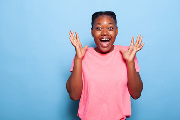 Portrait of excited african american teenager smiling at camera laughing while standing in studio with blue background. Young woman having energy making celebration facial expressions