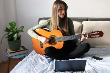 Young woman playing guitar.
Attractive young woman sits on the bed, plays acoustic guitar and reading notes on laptop.