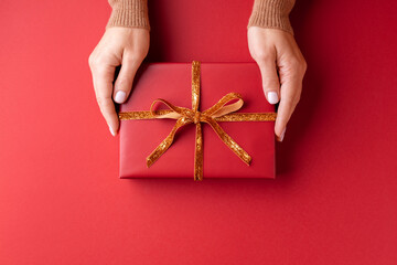 Female's hands holding red gift box with golden ribbon on red background.