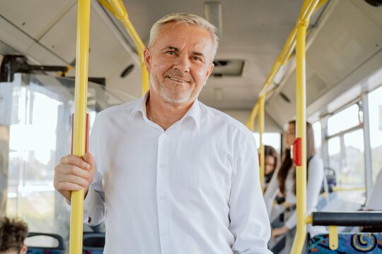 Grey-haired Elderly Man Dressed In Smart White Shirt Stands In Middle Of Bus, He Takes Public Transport To Work, Guy Is Holding Onto Railing Next To Door Opening Button, He Smiles At The Camera