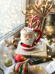 Cozy home Christmas atmosphere, window view mug of hot tea on windowsill, evening at home, holiday mood. Christmas decor with kraft house. Selective focus, blurred backdrop, vintage postcards