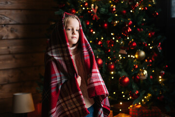 Portrait of pretty little blonde curly child girl wrapped in plaid looking at camera standing by decorated Christmas tree with bright festive lights. Happy kid came in morning to get presents at home.