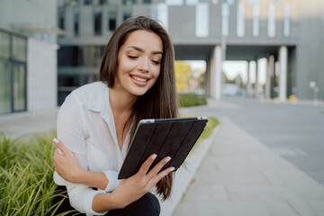 Break from office, smartly dressed girl sits on concrete outside in front of glass modern corporate building, ornamental plants around her, woman is holding a tablet, watching videos, series