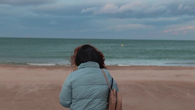 Woman with blue jacket observing a beach at sunset in winter